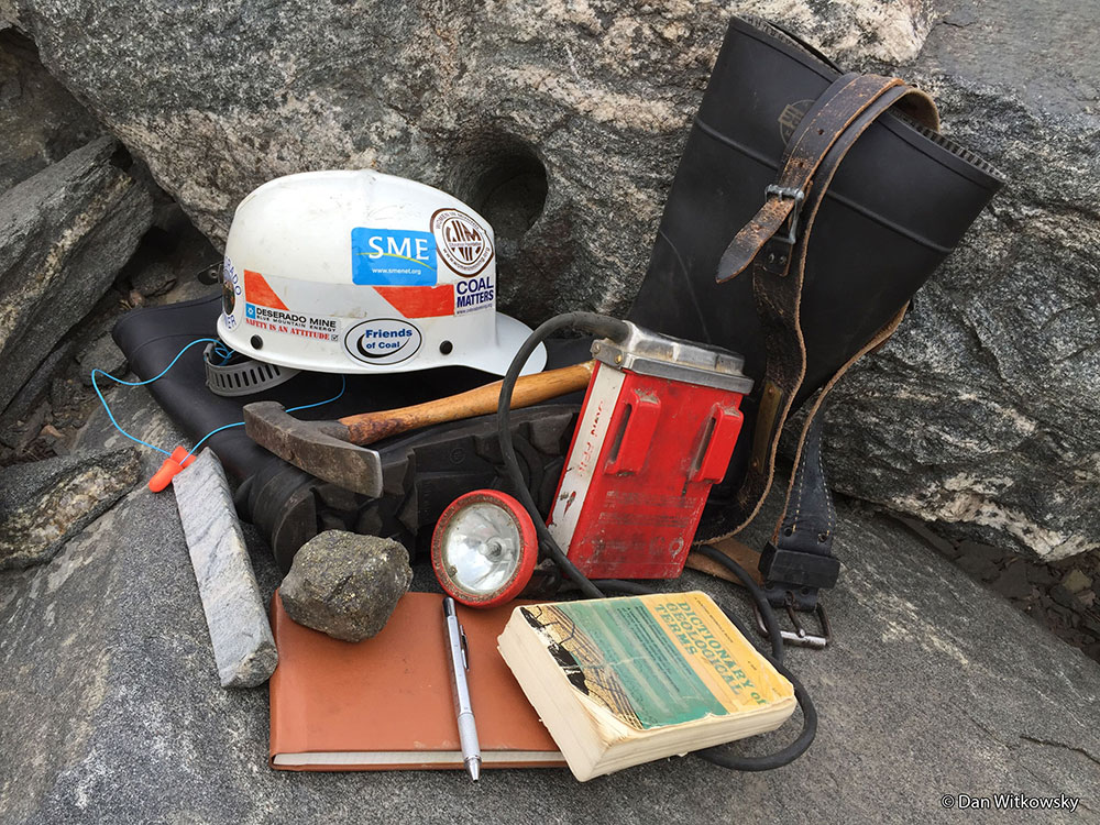 Honorable mention photo by Dan Witkowsky: A collection of mining tools, reference books, notebooks, hard hat, and boots, on a natural stone background